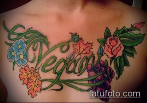 Фото тату: надпись "Vegan" в окружении винограда, листьев и цветов, выполненная на груди. Детализированный рисунок татуировки.