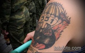 Фото тату на плече: изображение парашютиста с черепом внутри парашюта, окруженного пламенем. Черно-красный рисунок с детальной проработкой.
