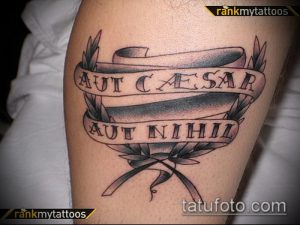 Фото татуировки с изображением свитка и латинской фразы 'Aut Caesar Aut Nihil' на мужской руке. Черно-серый рисунок выполнен в традиционном стиле.