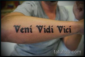 Фото татуировки с надписью 'Veni Vidi Vici' на мужском предплечье. Черные буквы четко выделяются на коже. Рисунок выполнен в классическом стиле.