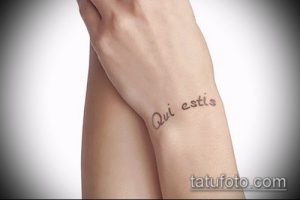 Фото тату на запястье женщины с надписью 'Qui estis'. Небольшой, изящный рисунок татуировки выполнен черными чернилами в каллиграфическом стиле.