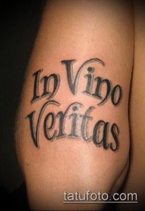 Фото тату: латинская фраза 'In Vino Veritas' на руке мужчины, выполненная черными чернилами в каллиграфическом стиле.