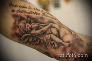 Фото тату: изображение ангела и латинская фраза на руке, выполненные в реалистичном стиле с использованием черно-серых оттенков.