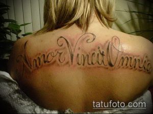 Фото тату на спине с латинской надписью «Amor Vincit Omnia» и декоративными завитушками. Черный цвет, классический стиль, женская татуировка.