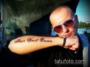 Фото тату на предплечье с латинской надписью «Amor Vincit Omnia». Классический стиль, черный цвет, мужская татуировка, вдохновляющий рисунок.