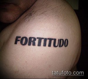 Фото тату на плече с латинской надписью 'FORTITUDO', выполненной черными чернилами. Классический и мужественный дизайн.