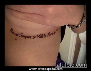 Фото тату с надписью 'Nunc et Semper te Valde Amabo' и маленьким сердечком на бедре. Тонкий шрифт, черные чернила, женственная татуировка.
