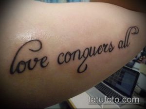 Фото тату на предплечье: надпись 'love conquers all' красивым курсивом. Черно-белый рисунок татуировки.