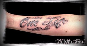 Фото тату надписи 'One Life' с декоративными завитушками на предплечье. Черно-серый рисунок, выполненный в каллиграфическом стиле.