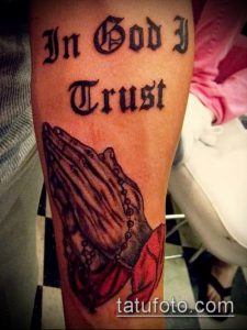 Фото татуировки на предплечье с надписью "In God I Trust" и изображением молящихся рук. Черно-белый рисунок татуировки с религиозным посланием.