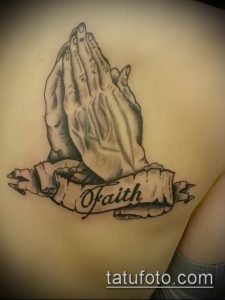 Фото тату молящихся рук с лентой и надписью "Of Faith", выполненное в реалистичном стиле с использованием черно-серых оттенков. Подходит для тех, кто хочет выразить свою веру.