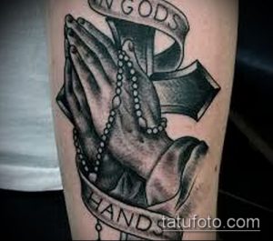 Фото тату молящихся рук с четками и лентой с надписью "In Gods Hands", выполненное в реалистичном стиле с использованием черно-серых оттенков.