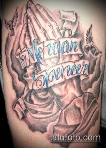 Фото тату молящихся рук с голубями и лентой с именами 'Morgan Spencer'. Детализированный рисунок татуировки на плече, выполненный в серо-голубой гамме.