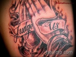 Фото тату молящихся рук в серо-черной гамме с лентой и надписью 'Judge Us'. Расположена на плече, детализированный рисунок татуировки.