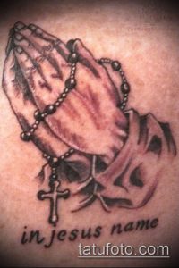 Фото тату на руке: молящиеся руки, четки, крест, надпись 'in Jesus name'. Черно-белый рисунок татуировки.