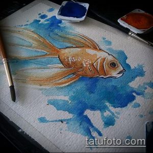 Акварельный эскиз татуировки золотой рыбки, плавающей в голубой воде, с использованием брызг и разводов краски.