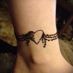 Фото мехенди на лодыжке: коричневый рисунок в виде браслета с сердцем и подвесками, выполненный в традиционном стиле.