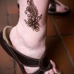 Фото тату мехенди на лодыжке с изображением цветка и растительных узоров, выполненных в черном цвете. Татуировка гармонично сочетается с сандалиями.