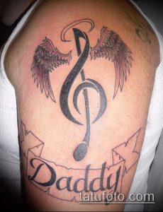 Фото тату на плече с изображением скрипичного ключа, крыльев и надписи 'Daddy'. Черно-белая татуировка с детальной прорисовкой.