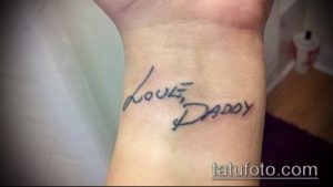 Фото тату надпись 'Love, Daddy' черными чернилами на запястье. Минималистичный рисунок татуировки, выполненный простым шрифтом.