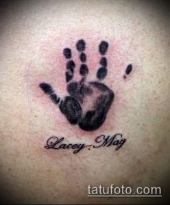 Фото тату: отпечаток детской ладошки с именем 'Lacey May' под ним, выполненный в черно-серых тонах на спине.