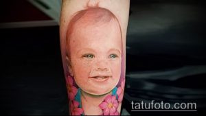 Фотография татуировки на руке, изображающей портрет улыбающегося ребенка, окруженного розовыми цветами, выполненной в ярких и нежных тонах.
