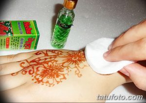 Фото процесса нанесения тату мехенди на руку, демонстрирующее работу мастера с аппликатором. Рядом с рукой расположены коробка с хной и бутылочка с маслом.