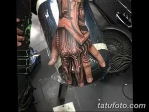 Фото тату механической руки на руке, выполненной в стиле киберпанк с использованием красных и серых оттенков. Детализированный рисунок с имитацией металлических пластин и проводов.