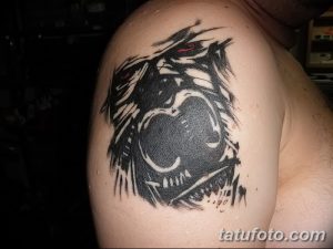 Черно-серая реалистичная татуировка головы разъяренной гориллы на плече мужчины. Красные глаза добавляют выразительности.