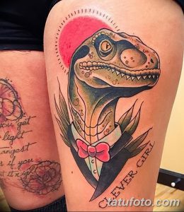Фото тату на бедре: яркий велоцираптор с бантиком и надписью "Clever Girl". Рисунок татуировки в стиле олдскул.