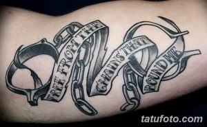 Фото тату: наручники, разорванные цепи и лента с надписью "Free from the chains that bound me" на руке.