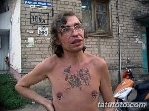 Фотография мужчины с татуировкой петуха на груди. Татуировка выполнена в серо-черных тонах, с детальной прорисовкой перьев и клюва. Мужчина в очках и с длинными волосами.