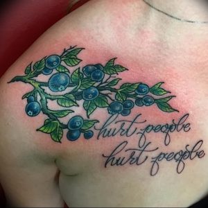 Фото тату с изображением ветки черники и надписи 'hurt people hurt people' на груди. Реалистичный рисунок, выполненный в ярких цветах.