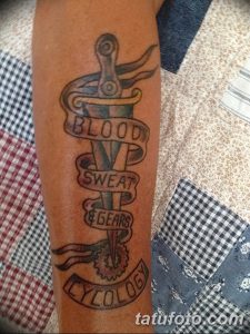 Фото татуировки на предплечье с вертикальными надписями "BLOOD, SWEAT & GEARS" и символами. Черно-серая татуировка в стиле традиционный.