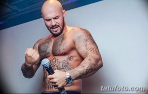 Фото мужчины с татуировками на руках и груди, выступающего с микрофоном. Татуировки выполнены в реалистичном стиле.