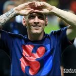 Лионель Месси с татуировками на руке, улыбается и смотрит в камеру. Фотография сделана на футбольном стадионе.