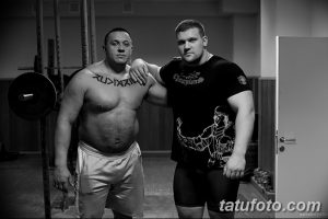 Фотография двух мужчин в спортзале, один из которых с татуировкой на груди. Черно-белое изображение подчеркивает мускулатуру и силу.