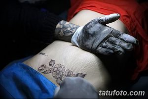 Фото тату: мастер наносит татуировку с изображением цветов на бок девушки. Видны рука мастера в перчатке и процесс нанесения рисунка.