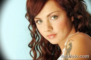Крупный план портрета девушки с татуировкой на плече. Девушка смотрит в камеру, ее волосы вьются. Татуировка выполнена в стиле трайбл.