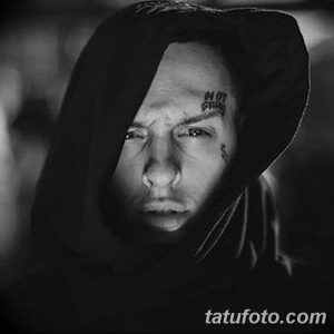 Черно-белый крупный план лица мужчины в капюшоне с татуировками под глазом и на лбу. Выражение лица серьезное и сосредоточенное.