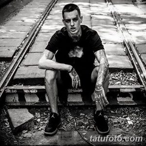 Черно-белое фото мужчины с обширными татуировками, сидящего на железнодорожных путях. Видны татуировки на руках, шее и груди.