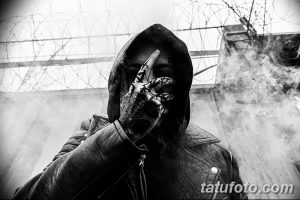 Черно-белое фото мужчины в капюшоне с татуировками на руках. Виден дым и проволочная сетка на фоне.