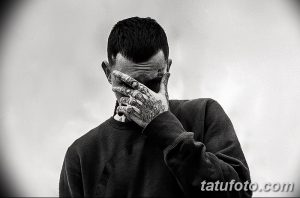 Черно-белое фото мужчины, закрывающего лицо рукой с татуировками. Видны татуировки на шее и руках.