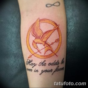 Фото тату сойки в круге с перекрещенными стрелами и надписью 'May the odds be ever in your favor' на предплечье.