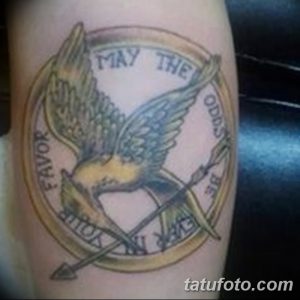 Татуировка сойки с перекрещенными стрелами в круге с надписью 'May the odds be ever in your favor' из 'Голодных игр'.