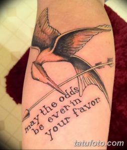 Фото татуировки сойки пересмешницы с цитатой 'may the odds be ever in your favor'. Черно-серый рисунок на предплечье.