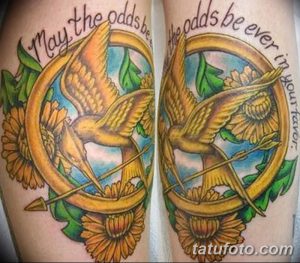 Фото татуировки сойки пересмешницы в круге с подписью 'May the odds be ever in your favor'. Красочный рисунок на руке.