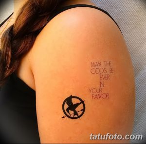 Фото тату: надпись 'May the odds be ever in your favor' и символ сокола в круге, выполненные на плече.