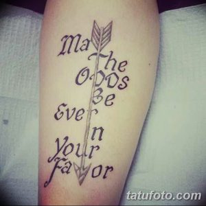 Фото тату: вертикальная надпись 'May the odds be ever in your favor' на руке, стилизованная под стрелу.