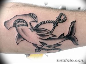 Фотография черно-белой татуировки на руке, изображающей молотоголовую акулу, обвивающую якорь. Татуировка выполнена в стиле олдскул с четкими линиями и деталями.
